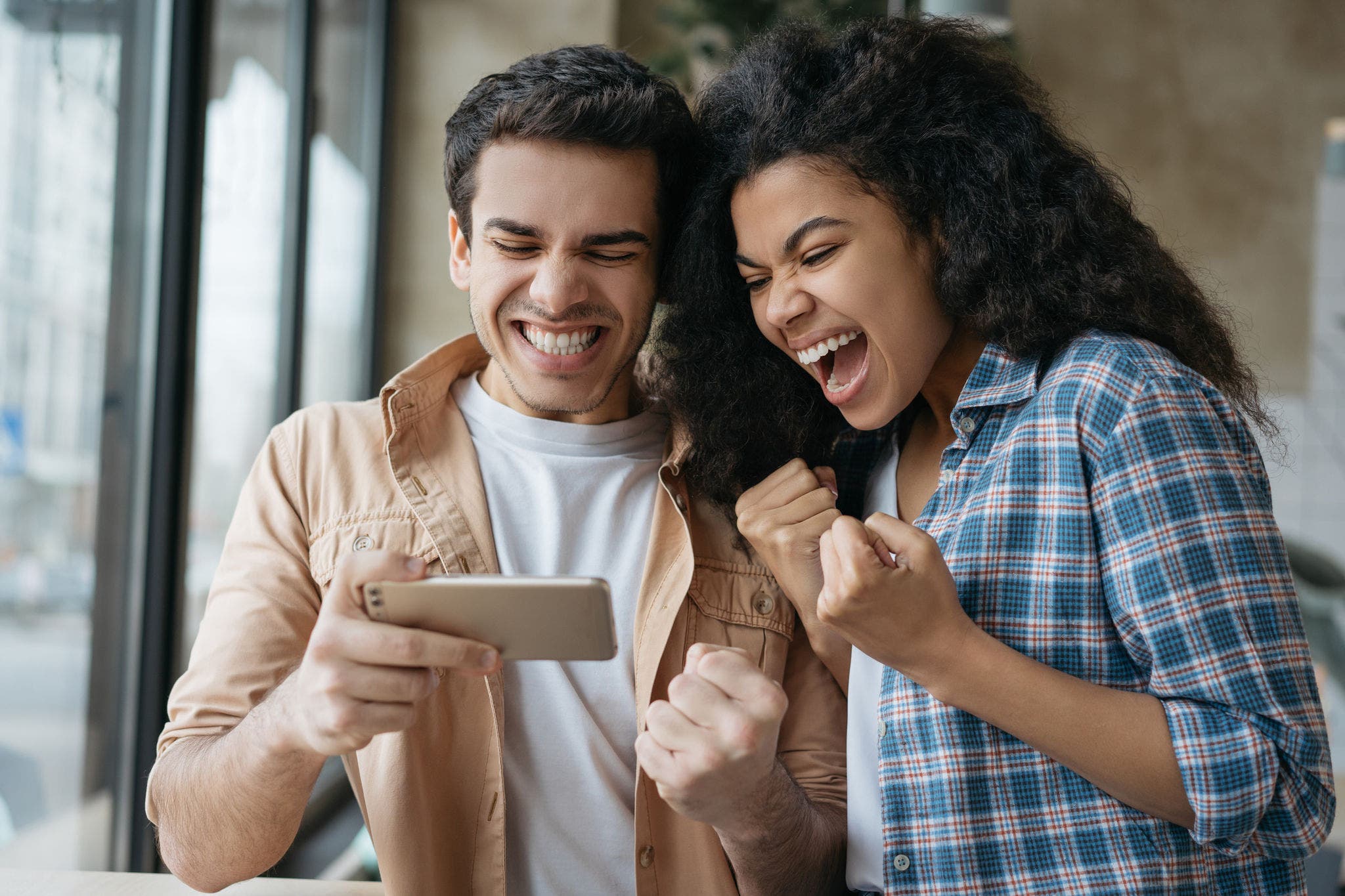 Young excited friends using smart phone, playing video games. Happy emotional couple celebration success. Portrait of overjoyed online lottery winners looking at digital screen. Sport betting concept
