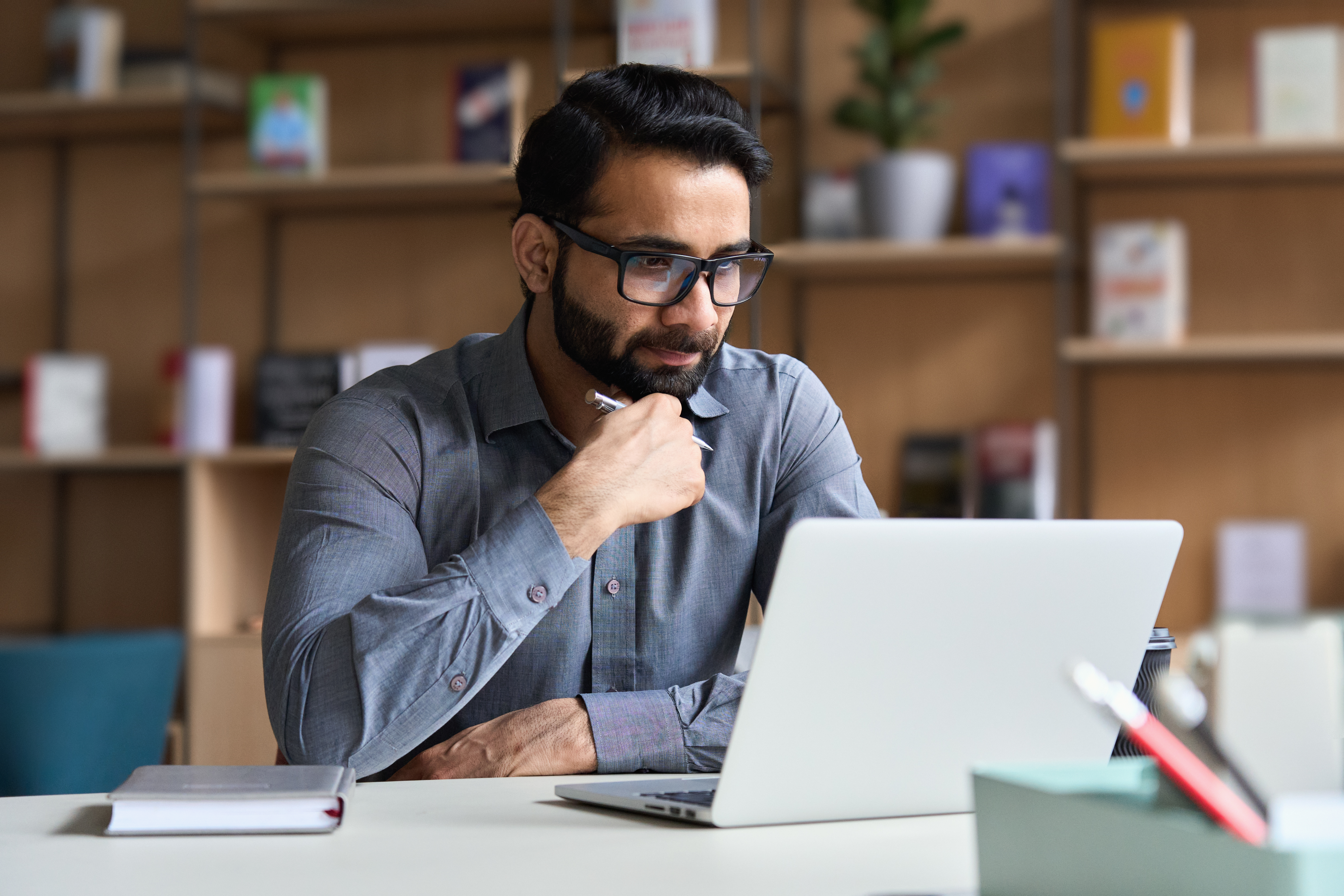 Young serious indian professional business man, focused ethnic male student wearing glasses working on laptop, remote studying using computer looking at screen watching seminar webinar at home office.